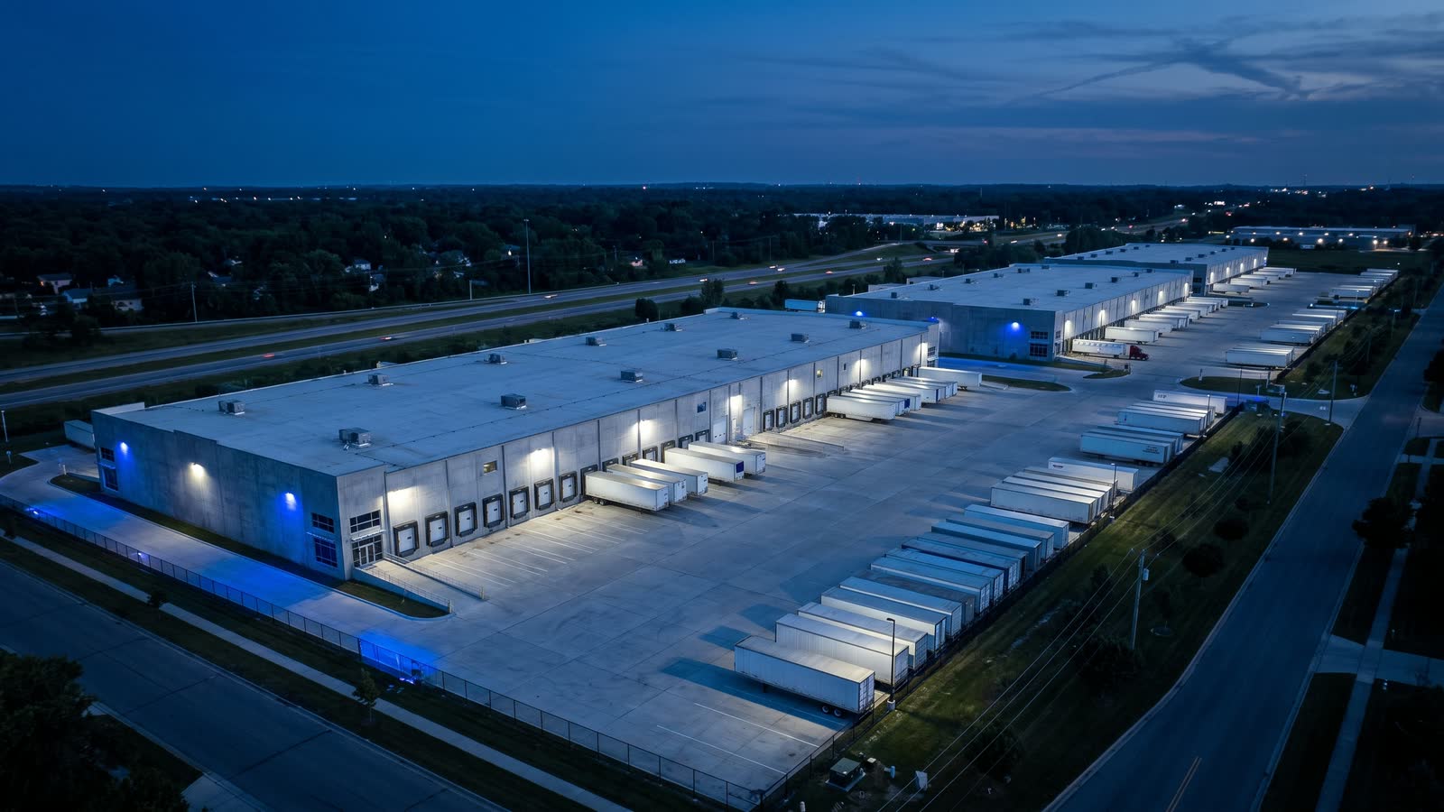 Blue-hour aerial of a multi-building industrial investment complex with rows of loading docks, a wide truck court and trailers parked along the bays.